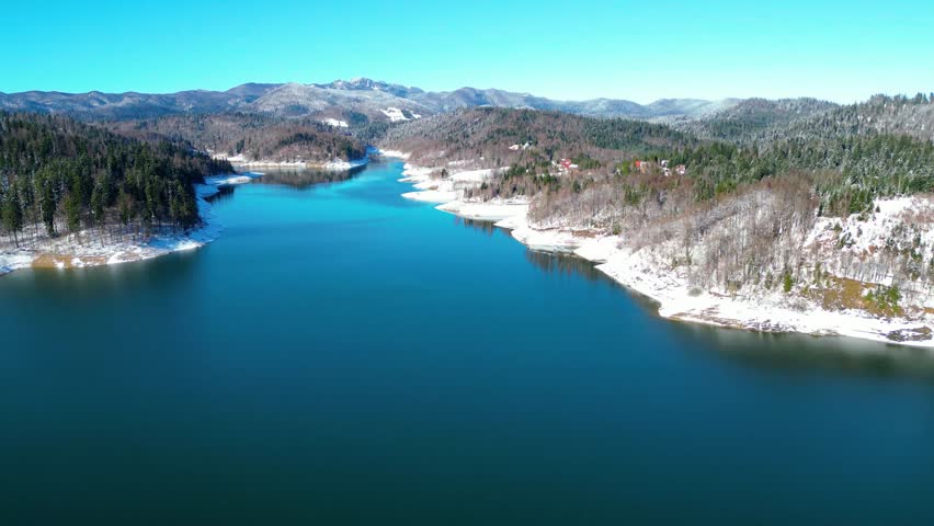 A stunning aerial view of Lokvarsko jezero, Croatia, blanketed in winter snow. The deep blue lake contrasts with pristine white forests under a clear sky.