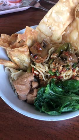  a bowl of chicken noodles with chopped chicken, chopped mushrooms, fried dumplings, chopped chilies and green vegetables on top. Video. Footage