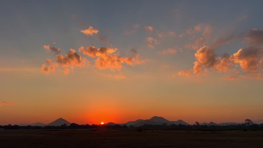 A serene landscape the sun setting behind distant mountains under a wide sky. Warm orange and golden tones illuminate scattered clouds, creating a peaceful scene. 