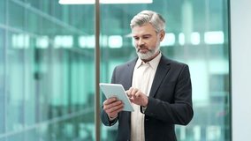 Confident mature businessman is using digital tablet while standing at workplace in modern glass business office. Entrepreneur in formal suit is chatting online, reading writing message, browsing web - Powered by Shutterstock - Get 15% off with code: PIKWIZARD15