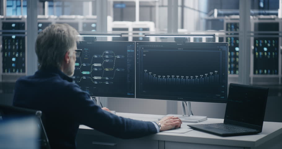 Senior Engineer Monitoring Data Flow on Dual Screens and Laptop. IT Specialist Working on Monitors Displaying Complex Network Diagrams and Analytics in Data Center. Network Security and Monitoring.