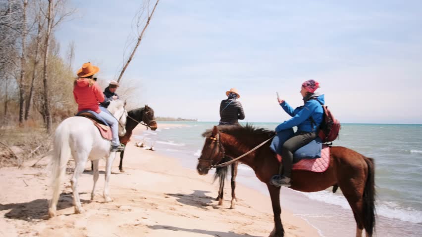 Group of people on horseback stop by the lake shore during an equestrian tour, taking photos and enjoying the nature. Sunny day, calm water, relaxed atmosphere.