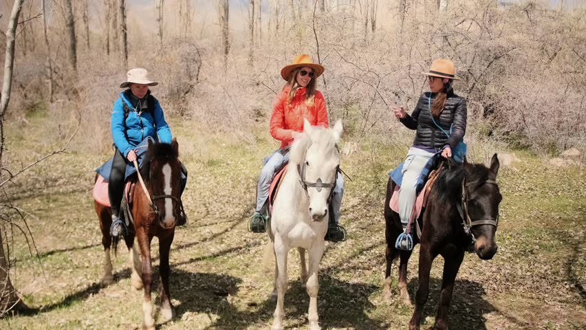 Three friends riding horses in a wooded area, stopping to take pictures of one another with their smartphones. Rural adventure and modern habits, slow tourism and connection to nature.