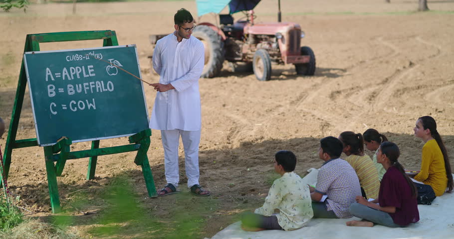 Indian young adult man tutor teach English asking questions to villager girl boy group outdoor rural area ground place. Happy teen age student team hold pencil sitting on mat look board learn alphabet outside open air class filed