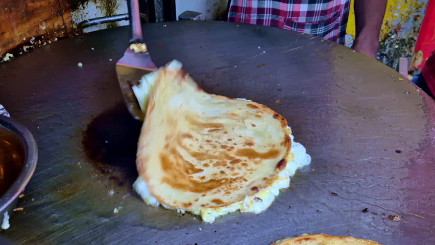 A close-up of a large street-food griddle with a freshly cooked eggroll paratha on one side, a dark heat mark at the center, and chopped vegetables visible in the background.