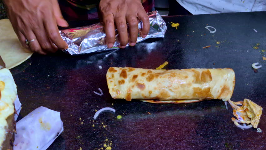 A street-food griddle scene showing a rolled eggroll paratha wrap resting on a hot surface, with scattered vegetable scraps and partially cooked flatbreads around it.