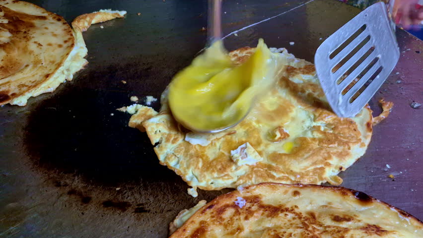 A close-up of freshly cooked eggroll on a hot flat griddle, being flipped with a metal spatula as butter or oil is spread, showing golden-brown layers and crisp edges.