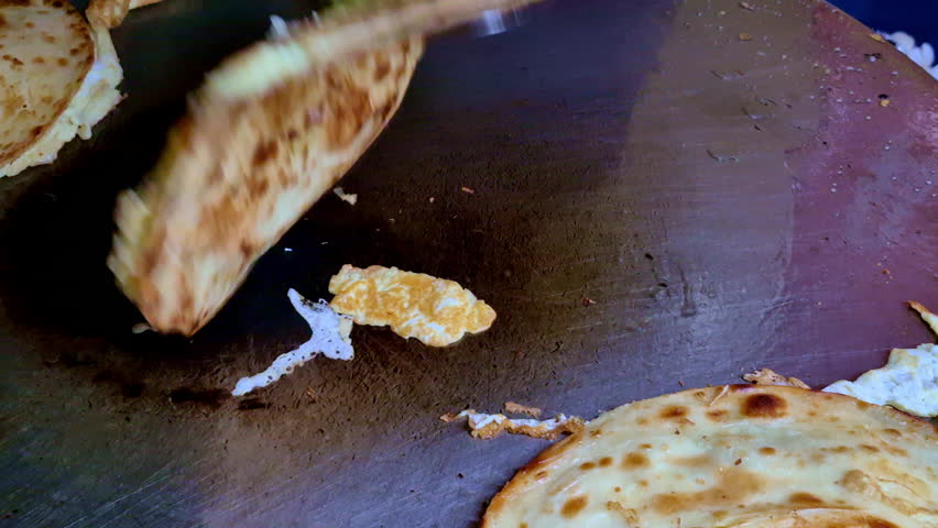 A close-up of multiple eggroll parathas cooking on a large flat griddle, showing evenly browned surfaces, flaky layers, and a metal spatula ready for flipping.