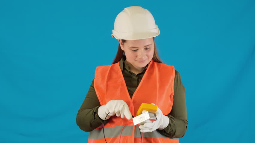 Positive female engineer holds multimeter showing thumbs up against blue. Woman builder in uniform expresses satisfaction with results