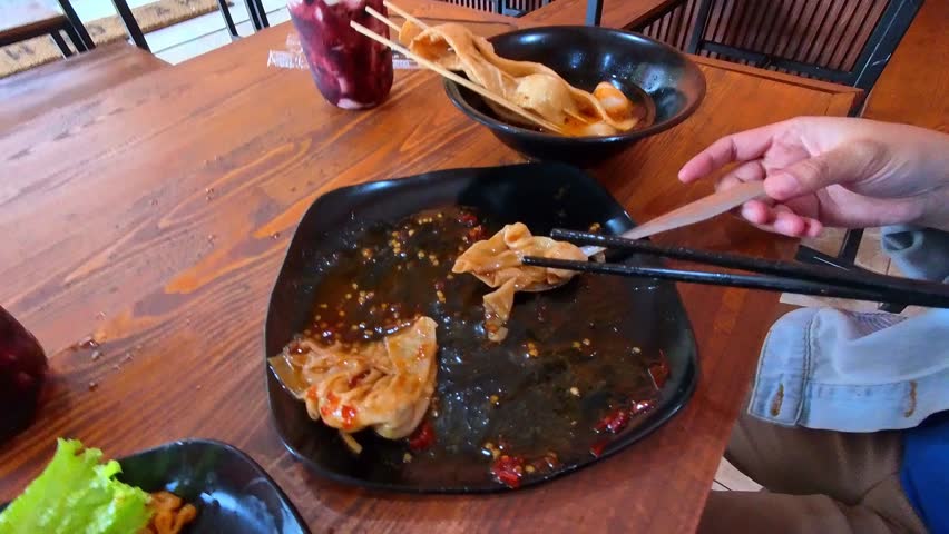 Overhead shot clip of eating spicy dumplings in a restaurant. Videography of Chinese food served on a black plate with chopsticks and a spoon in 4K HD resolution.