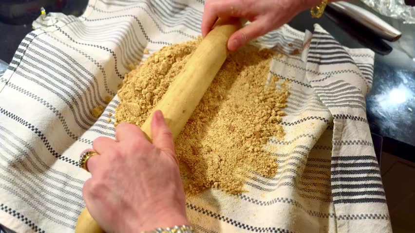 woman in kitchen crushing biscuit into crumbs for baking with wooden rolling pin