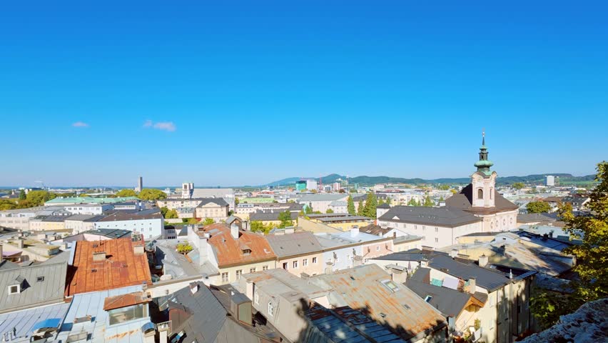 SALZBURG, AUSTRIA – OCTOBER 3, 2025: A high-angle panoramic view of the city rooftops featuring historic buildings and church spires under a clear blue sky. The cityscape showcases traditional European architecture and urban development stretching toward the horizon.