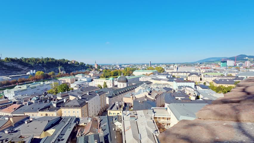 SALZBURG, AUSTRIA – OCTOBER 3, 2025: A high-angle panoramic view overlooking the historic city center with its traditional rooftops, church spires, and the river flowing through the valley. The scene captures the dense urban architecture and surrounding green hills under a clear blue sky.