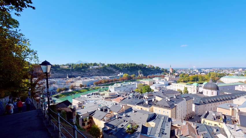 SALZBURG, AUSTRIA – OCTOBER 3, 2025: A high-angle panoramic view overlooking the historic city center with its traditional rooftops, church spires, and a large dome. The scene captures the dense urban architecture and surrounding green hills under a clear blue sky.