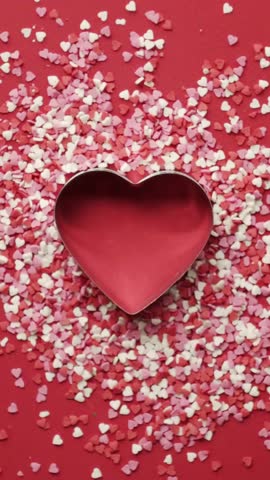 A top-down view of a red surface covered in a multitude of small, heart-shaped sprinkles in shades of red, pink, and white. A metallic heart-shaped cookie cutter is placed in the center, and a person