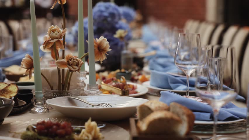 Elegant banquet table with floral arrangements, candles, glassware and blue napkins prepared for festive celebration, refined restaurant interior setup for wedding, christening or family event