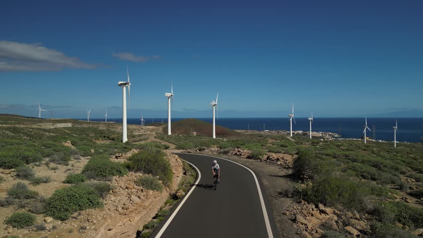 A lone cyclist enjoys a challenging ride on a winding road through an arid, coastal landscape under a clear blue sky, passing modern wind turbines with the vast ocean in the background