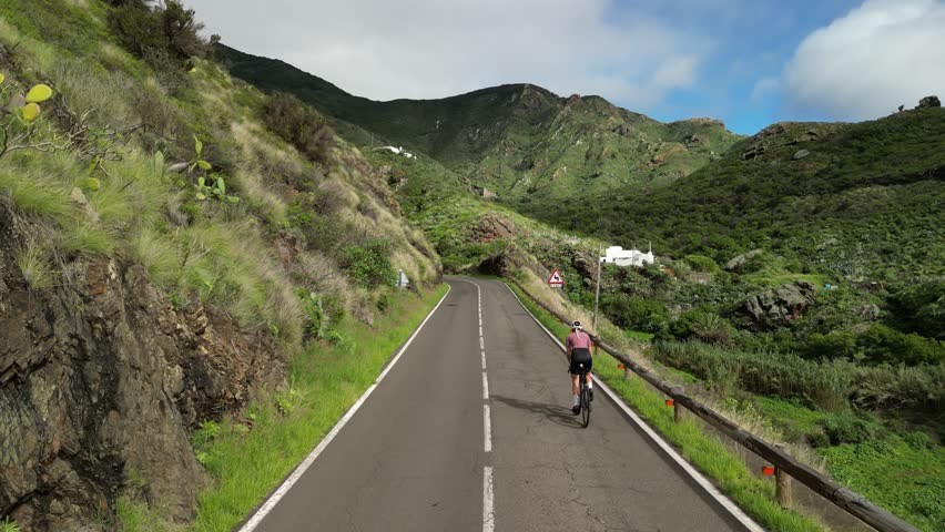 A cyclist pedals along a winding mountain road, surrounded by lush green hills under a cloudy sky. This scenic route offers a challenging yet rewarding outdoor adventure through nature