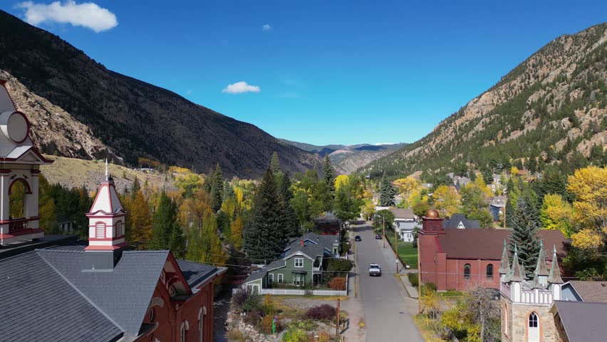 Cinematic aerial drone footage slowly rising over Georgetown, Colorado, showcasing a small mountain town surrounded by trees with changing autumn leaves. Smooth motion captures charming streets, colorful foliage, and the scenic mountainous environment. Ideal for travel, seasonal, small-town, lifestyle, and background visuals.