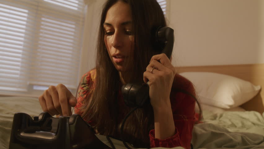 Retro woman in red dress excitedly calls friend on old rotarty telephone and rolls over in bed - close up