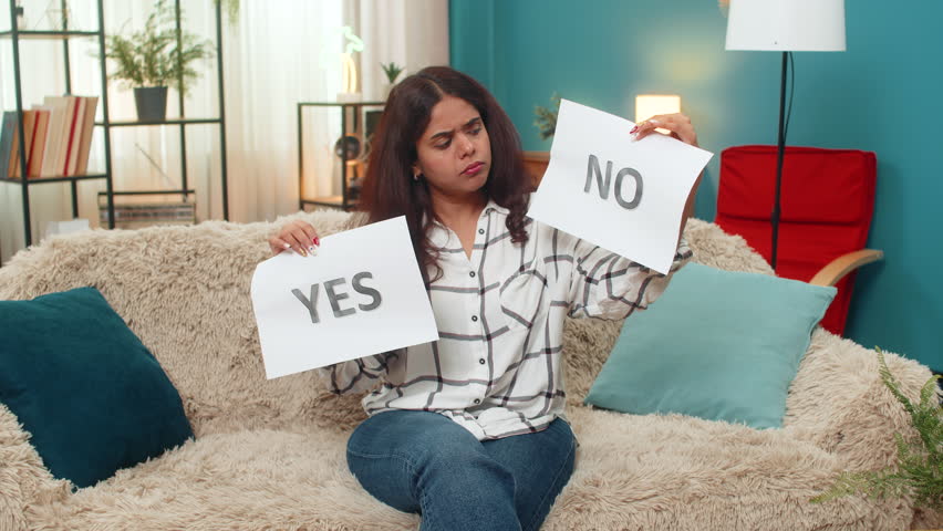 Indian woman at home holds yes and no papers hesitating choosing feeling stressed confused. Arabian girl on sofa looks at camera seeking advice showing doubt decision dilemma anxious mood alert tense.