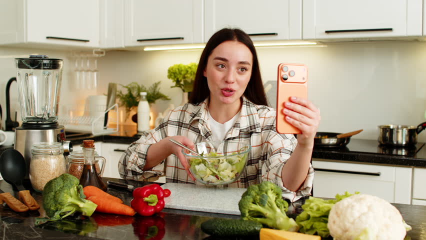 Young woman films herself on mobile phone, sharing keto diet tips and nutrition advice with followers. Blogger presents salad bowl, explains low carb ingredients, meal prep ideas, and routine at home.
