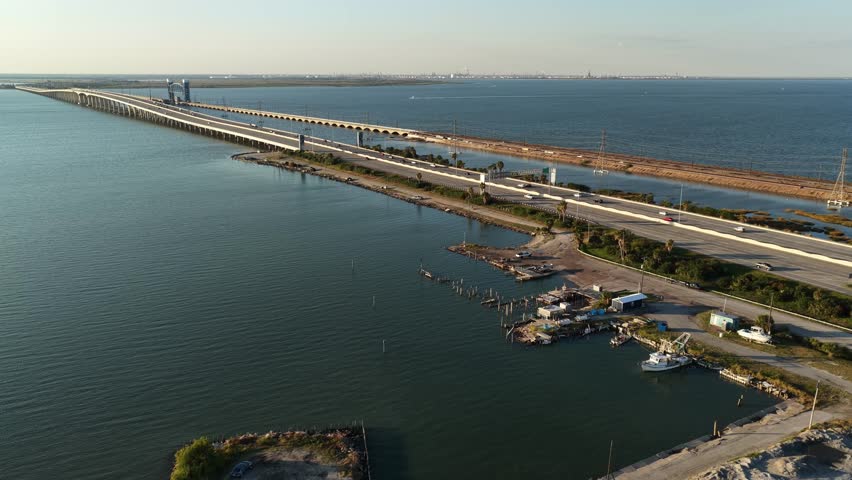 Houston, Texas, United States – November 2025.
High-altitude aerial view of a major roadway bridge spanning coastal waters in Houston. The drone performs a smooth forward movement with slight lateral alignment, capturing cars, trucks, and urban transportation flow along the bridge, with industrial elements and distant skyline visible on the horizon. Editorial use only.