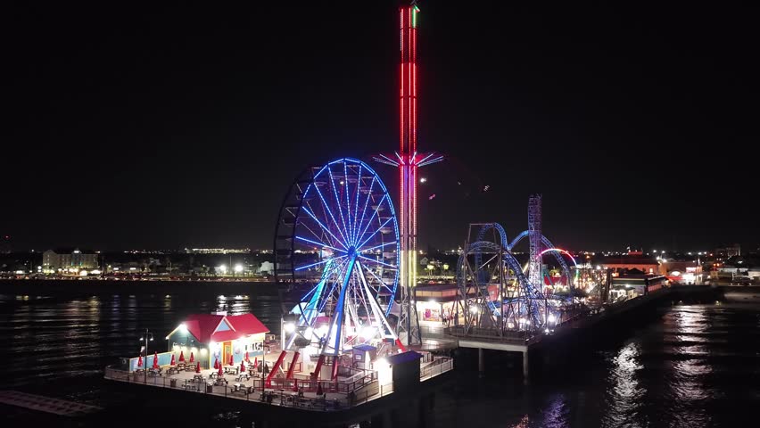 Galveston Island, Texas, United States – November 2024:
Night aerial drone view of the Galveston Island Historic Pleasure Pier featuring a brightly illuminated Ferris wheel, vertical drop tower, and roller coaster extending over the Gulf of Mexico. The video captures reflections on the water and the vibrant nighttime atmosphere of this popular coastal entertainment destination.