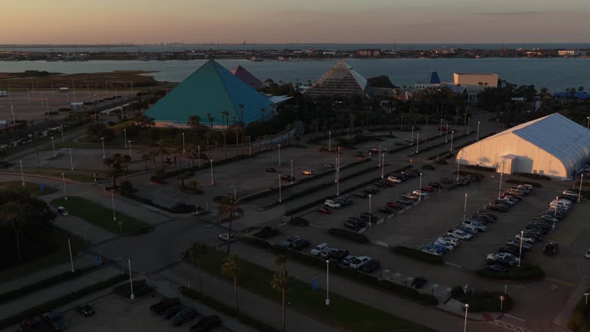 Galveston Island, Texas, United States – November 2024:
Aerial drone footage with a slow tilt-down movement from the horizon toward the glass pyramid, capturing the transition from sunset sky to architectural details and palm-lined walkways.