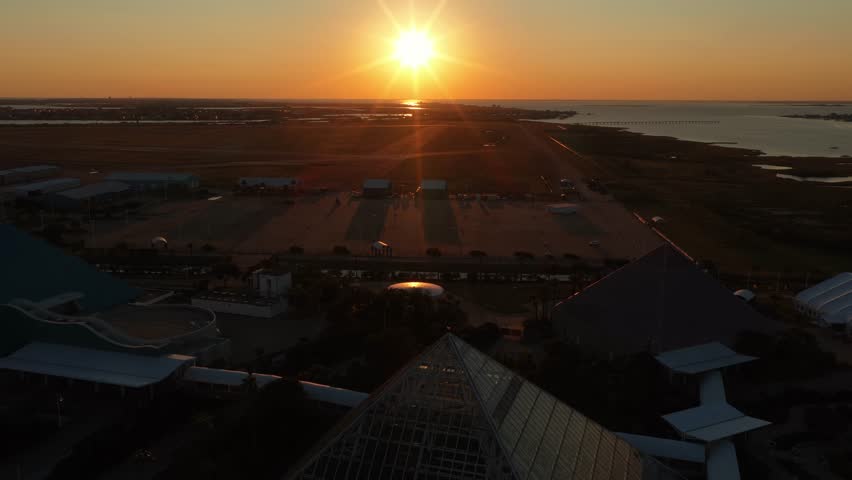 Galveston Island, Texas, United States – November 2024:
Aerial drone footage at sunset showing a smooth forward movement toward the iconic glass pyramid at Moody Gardens. The shot reveals the surrounding parking areas, palm trees, and coastal wetlands with warm golden-hour light reflecting across the landscape.