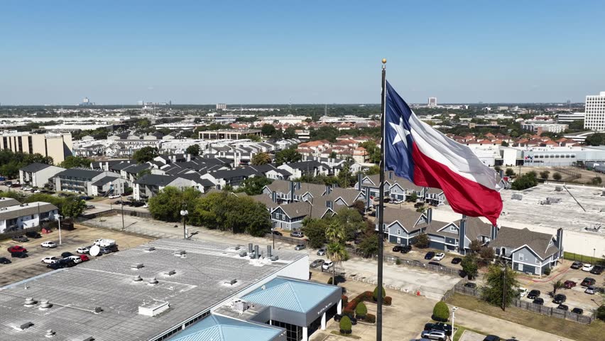 Houston, Texas, United States – November 2025:
Aerial drone view of the Texas state flag waving prominently above an urban residential neighborhood in Houston. The footage shows rows of apartment buildings, streets, parking lots, and rooftops under clear blue skies, highlighting everyday city life and suburban development in one of the largest metropolitan areas in the United States.