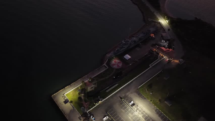 Galveston Island, Texas, United States – November 2024:
Night aerial drone footage showing a slow forward movement over a coastal parking area near the waterfront, revealing parked vehicles, illuminated streetlights, and calm water in the background.