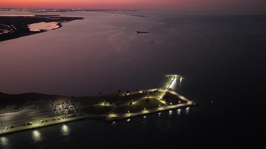 Galveston Island, Texas, United States – November 2024:
Nighttime aerial drone video with a gradual pull-back motion, revealing a coastal park area connected to a pier and surrounded by calm ocean waters.