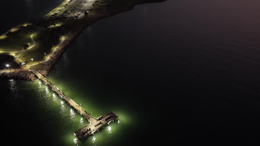 Galveston Island, Texas, United States – November 2024:
Night aerial footage featuring a descending drone movement over a coastal park and pier, with light reflections visible on the surrounding water.