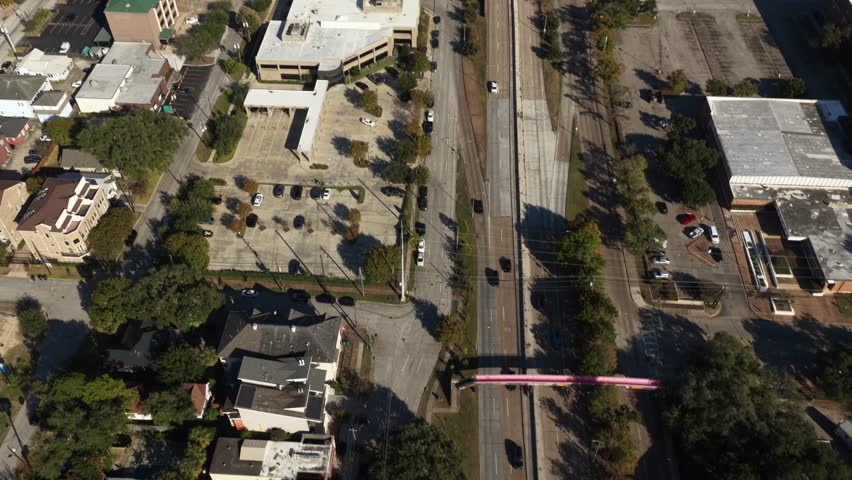 Houston, Texas, United States – November 2025:
Top-down aerial drone view of an urban neighborhood in Houston showing residential buildings, tree-lined streets, and a multi-lane roadway with moving vehicles. A pedestrian bridge crosses the avenue, highlighting everyday city infrastructure and urban mobility from a vertical perspective