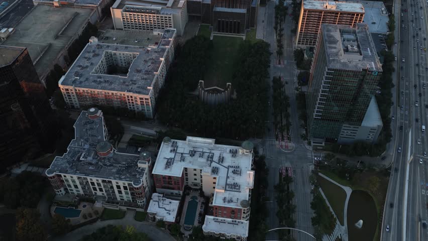 Houston, United States, on October 28, 2024.
The image shows a geometric city layout composed of modern residential and office buildings surrounding a rectangular green park area densely covered with trees. At the center of the park, a distinctive architectural structure is visible, partially enclosed by vegetation, creating a strong contrast between natural space and surrounding urban development.
The drone flies at a high altitude, offering a top-down perspective that clearly reveals the relationship between the park, nearby streets, and adjacent high-rise buildings. Wide avenues, intersections, and traffic lanes frame the green area, highlighting Houston’s urban planning and large-scale infrastructure. The surrounding architecture features a mix of contemporary glass towers and mid-rise residential complexes, typical of downtown and inner-city Houston neighborhoods.
Natural daylight and soft shadows suggest late afternoon lighting, enhancing textures on rooftops, streets, and treetops. The aerial viewpoint provides a clear understanding of spatial organization, land use, and the integration of green public spaces within a dense metropolitan environment.
This editorial image represents themes such as urban development, city planning, public parks, architecture, and modern American city life. It is suitable for editorial use related to travel, geography, infrastructure, urban growth, and city landscapes in the United States.