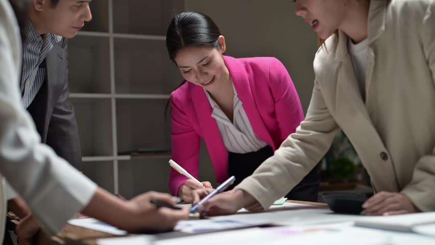 A woman in a pink jacket is writing on a piece of paper. She is surrounded by other people