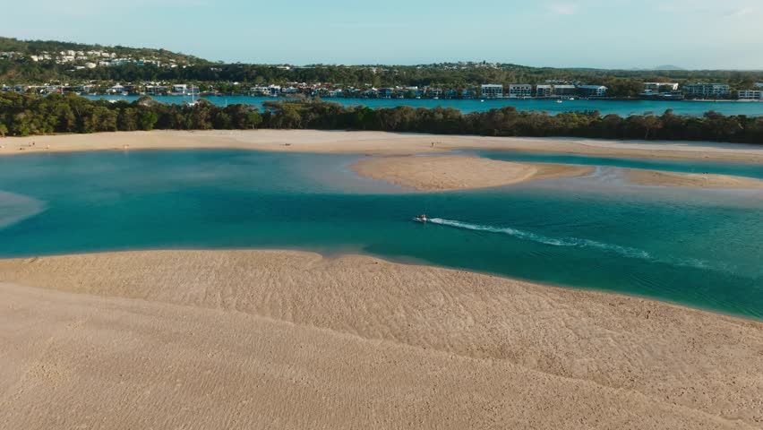 High angle drone orbits boat driving in shallow turquoise water near sandy beach, up river, Noosa Queensland Australia