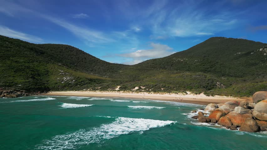 Aerial coastal approach to turquoise surf breaking near rocky shore beneath forested hills, Wilsons Promontory Victoria Australia