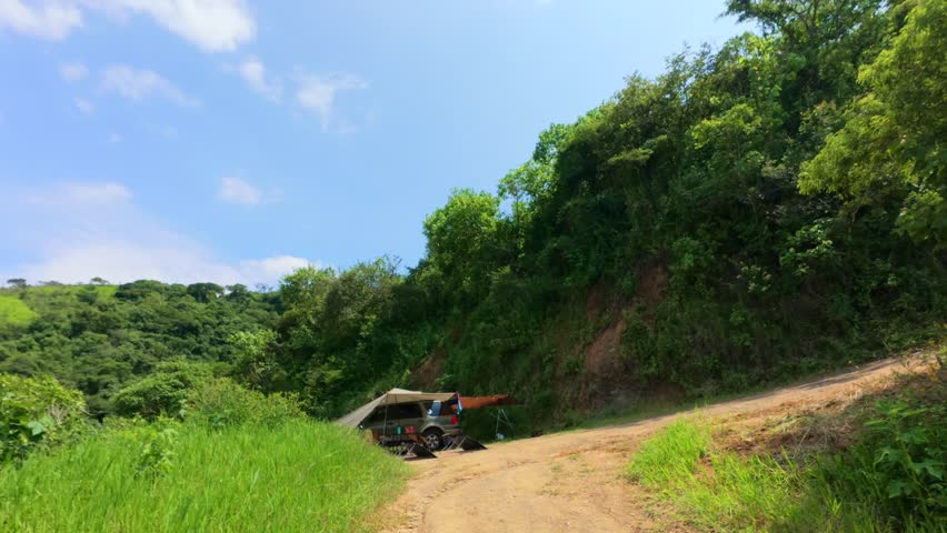Couple in swimwear walks on dirt road toward SUV camper surrounded by green hills at Presa El Carrizo, Mexico