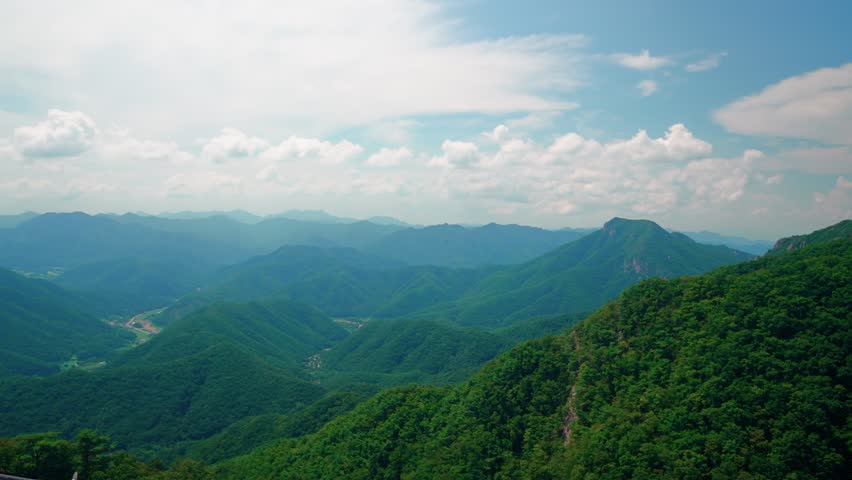 Wide angle landscape showing rolling green hills and mountain peaks of Daedunsan Provincial Park under cloudy blue sky from high viewpoint with traditional rooftop in South Korea