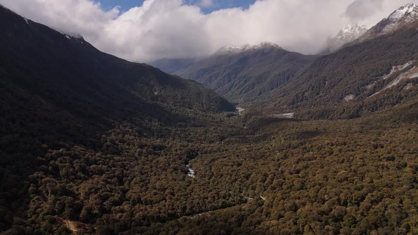 Aerial wide shot of Fiordland National Park with green mountains on sunny day. Clouds behind at Milford Sound in New Zealand.