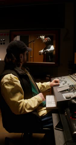 The sound engineer controls the recording process of the song in the studio. The rapper sings in the room behind the glass
