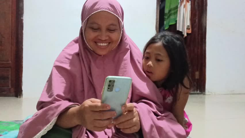 An Indonesian Muslim mother in a pink hijab sits with her young daughter, smiling and laughing together while looking at a smartphone screen. A heartwarming family moment in a simple home setting.