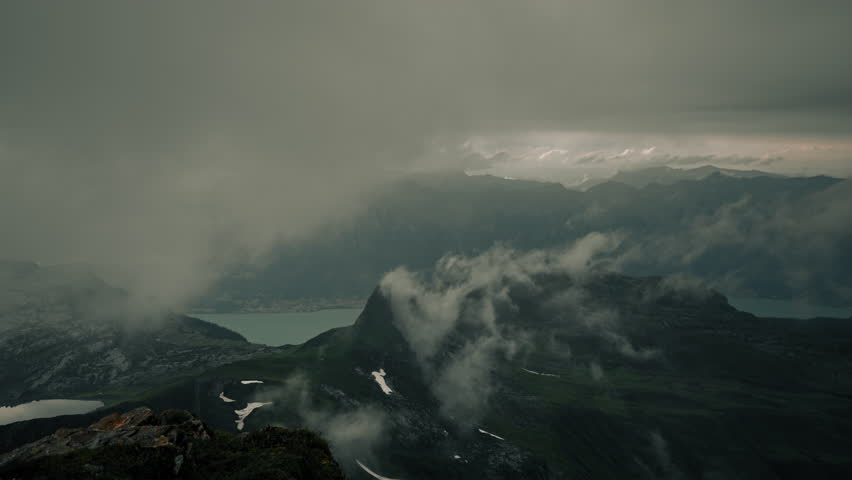 Epic wide view of a dramatic alpine mountain panorama with heavy clouds rolling over green ridges and a distant turquoise lake revealed beneath the stormy Swiss Alps sky
