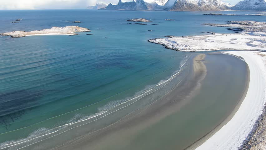 Aerial view of a snow-covered white sand beach near Fredvang and Ramberg, where the winter Lofoten landscape glows under a steel-blue sky in crisp Arctic light.