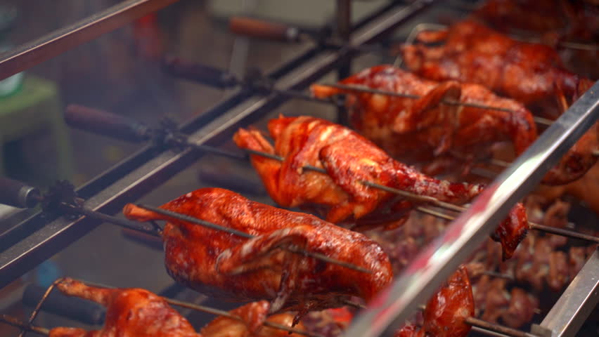 A high-quality close-up shot of multiple whole ducks with golden, crispy skin rotating slowly on a mechanical rotisserie spit. Glistening fat, caramelized texture, and subtle wisps of smoke.