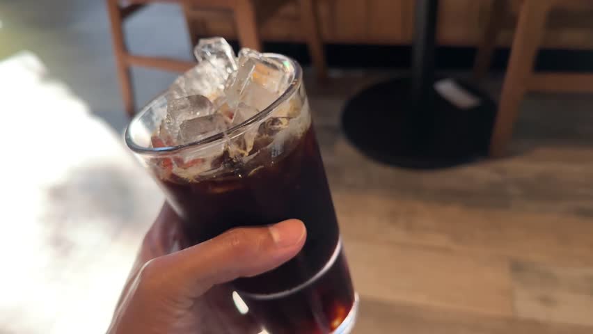 Close-up of Hand Holding Tall Glass of Refreshing Iced Black Coffee Americano in Modern Cafe Lifestyle Background