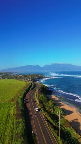 An aerial shot captures a scenic coastal road in Maui, Hawaii, winding alongside the Pacific Ocean with waves crashing against the shore.