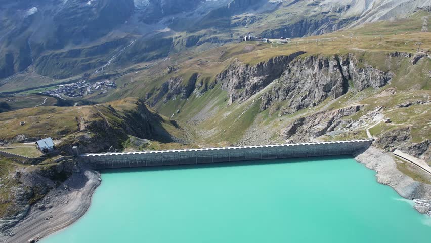 Aerial view of the Diga del Goillet dam holding back a turquoise lake, contrasting with the rugged mountain landscape, Diga del Goillet, Valle d
